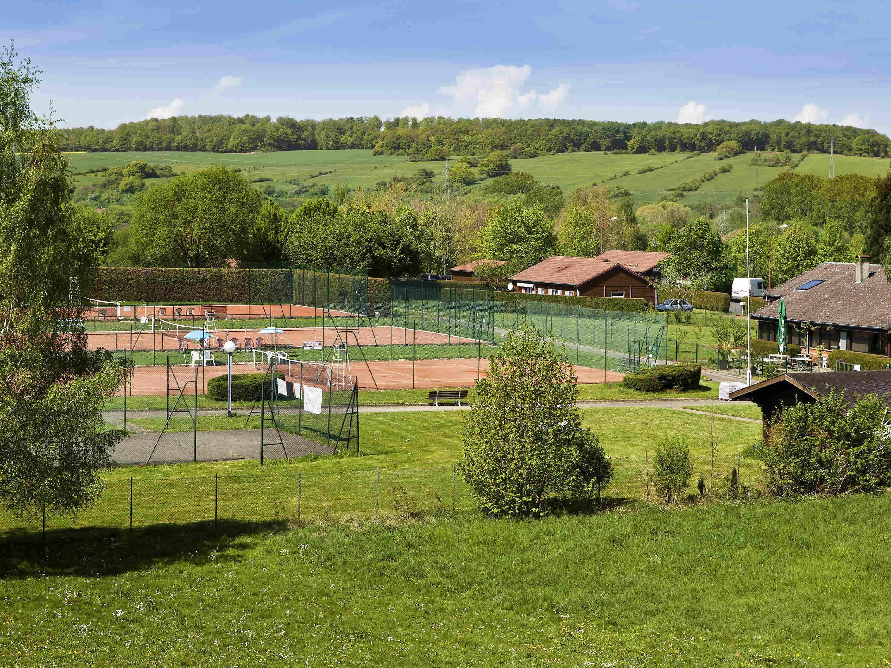 Una cancha de tenis rodeada de campos verdes y árboles, con un edificio al fondo.
