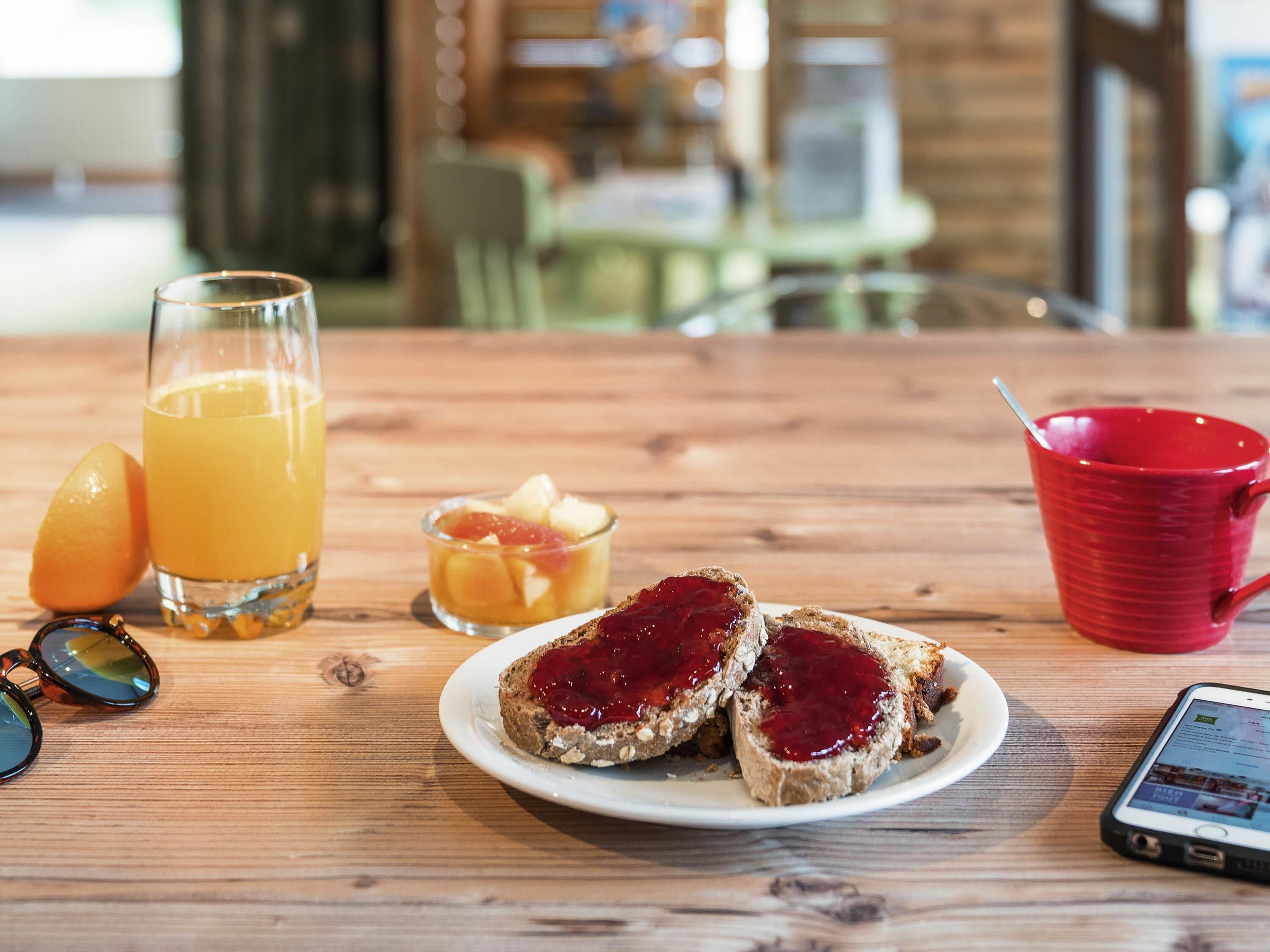 Ett träbord med frukosttillbehör, inklusive rostat bröd med sylt, ett glas apelsinjuice, en röd mugg och en smartphone.