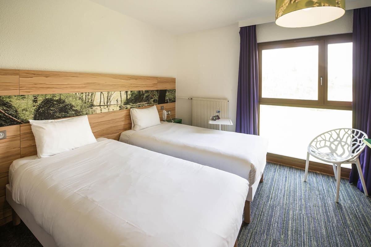 A hotel room with two beds, a radiator, a window with curtains, and a white chair.