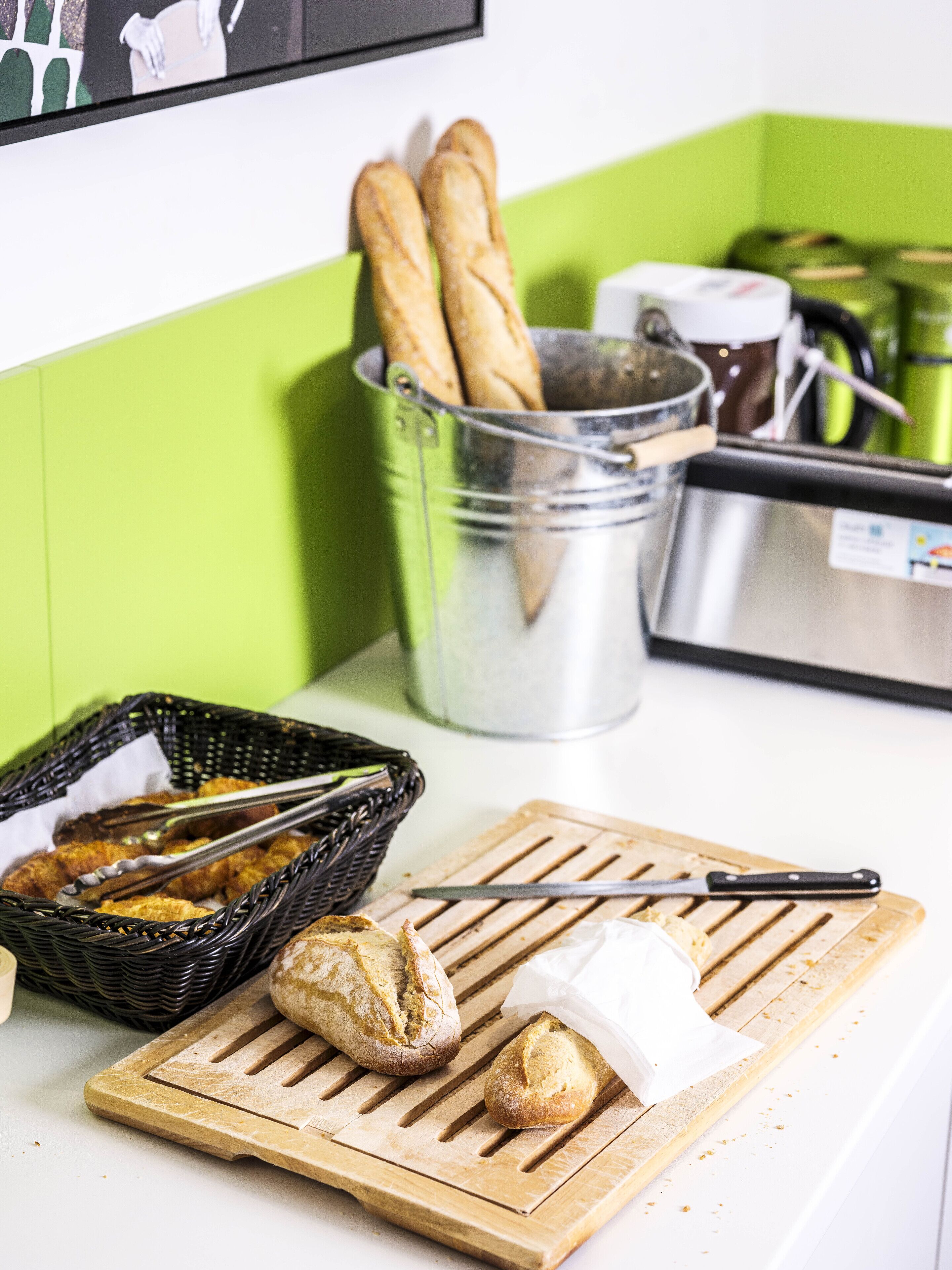A kitchen counter with a cutting board, tongs, a basket of bread, a metal container, and a green wall.