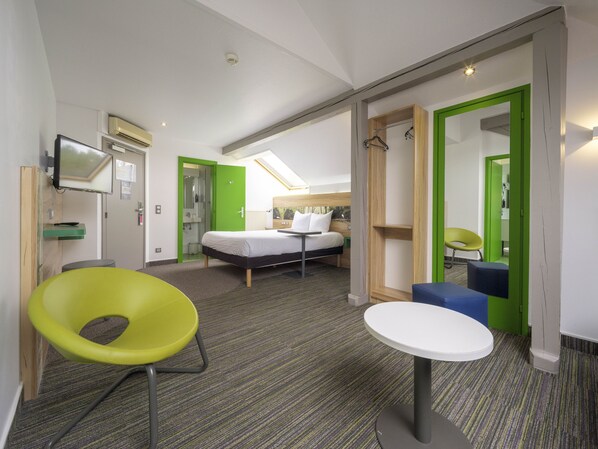 Habitación de hotel con cama, escritorio, silla y una pequeña mesa. Se observa un espejo, una ventana y una puerta que da a otra habitación.