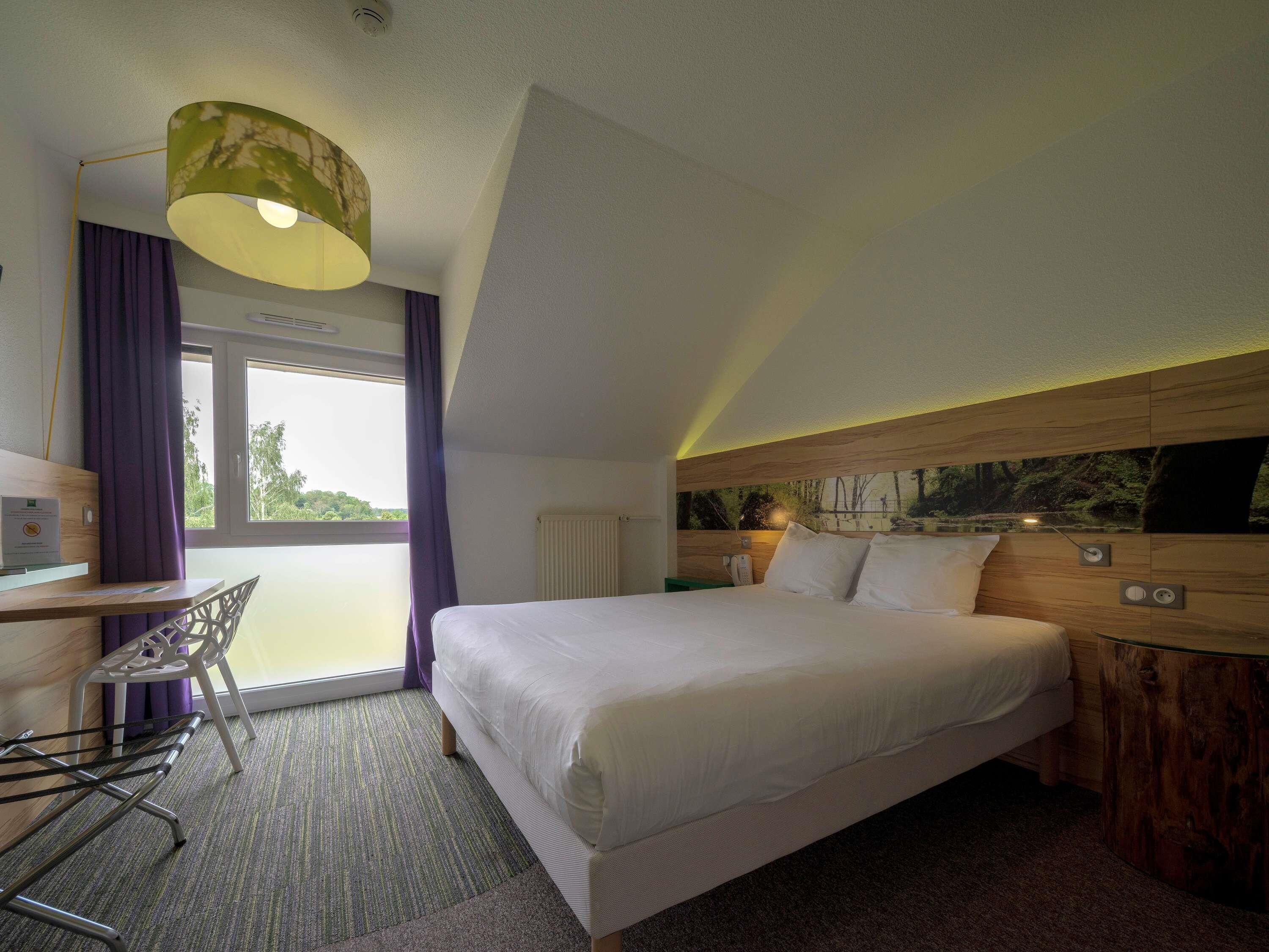 Une chambre d’hôtel comprenant un lit, un bureau, une chaise, une fenêtre avec des rideaux, un radiateur et un téléviseur fixé au mur.
