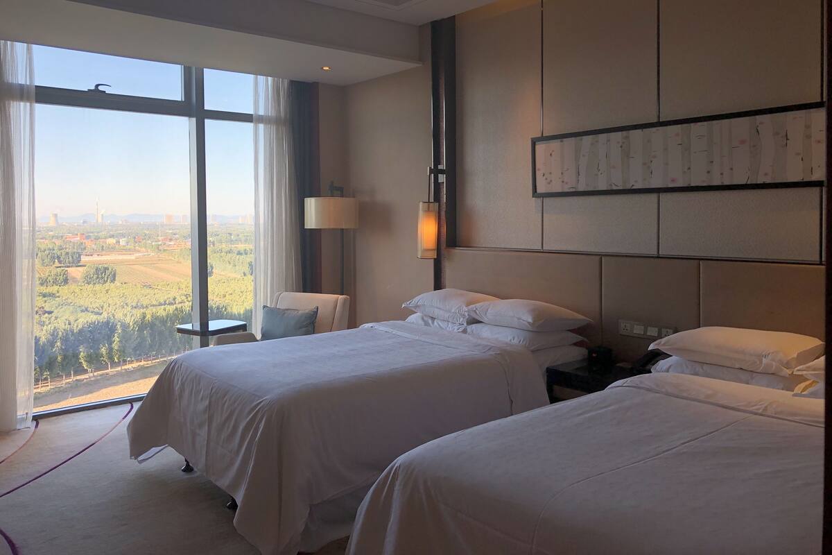 A hotel room with two beds, a large window with a view of a landscape, a bedside table, and a wall-mounted lamp.