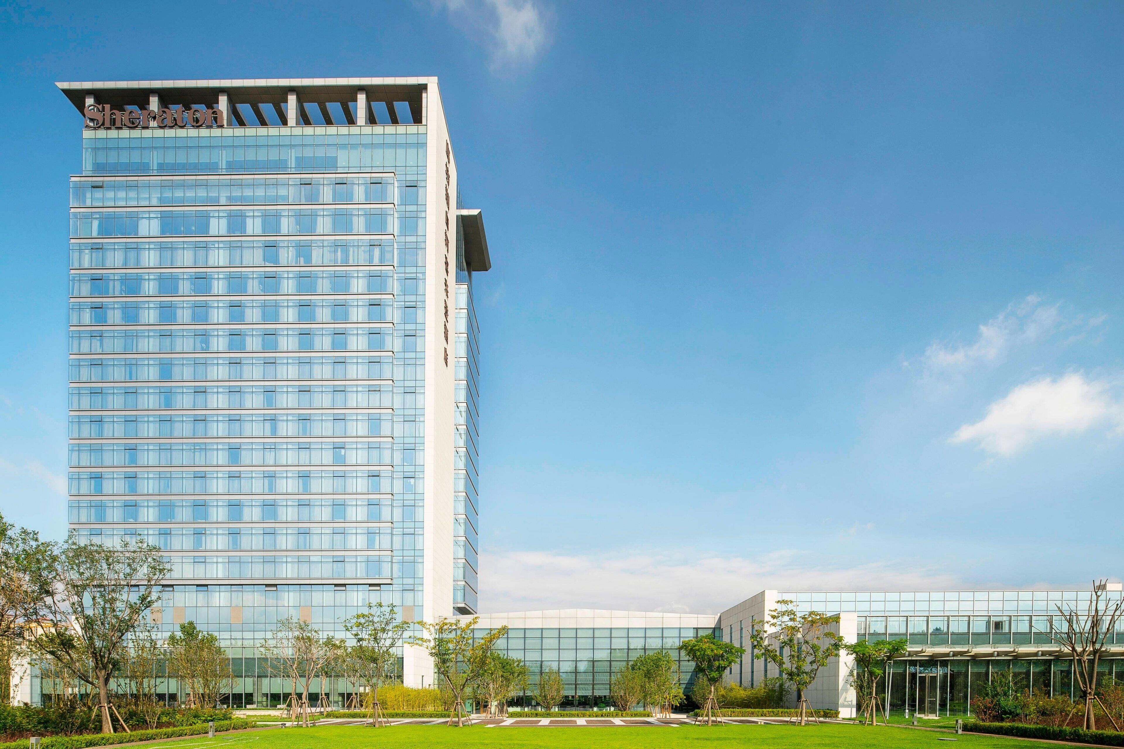A modern high-rise hotel with a glass facade, a well-maintained lawn, and a clear blue sky.