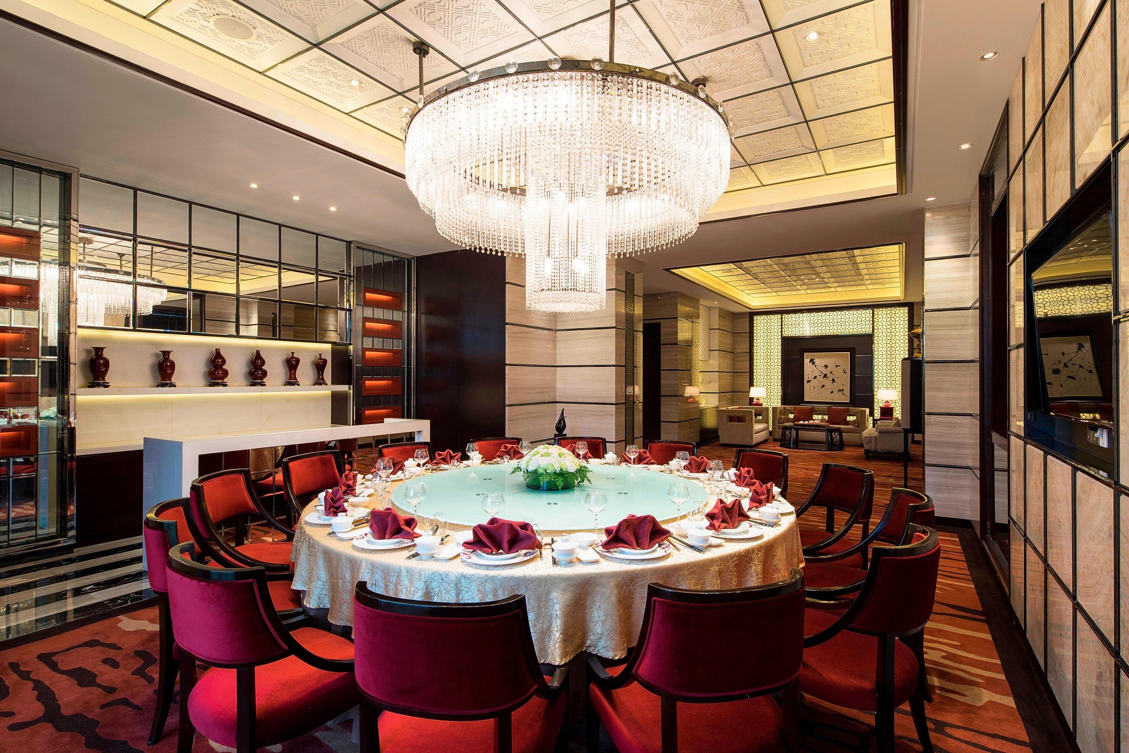 A luxurious dining room with a large chandelier, round table, and red chairs.