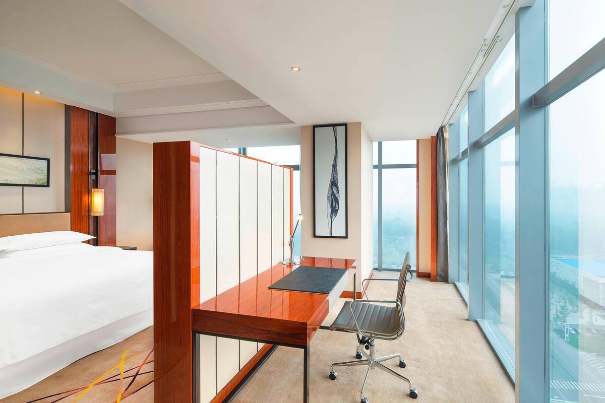 A modern hotel room with a large bed, a wooden desk, a chair, and a large window offering a city view.