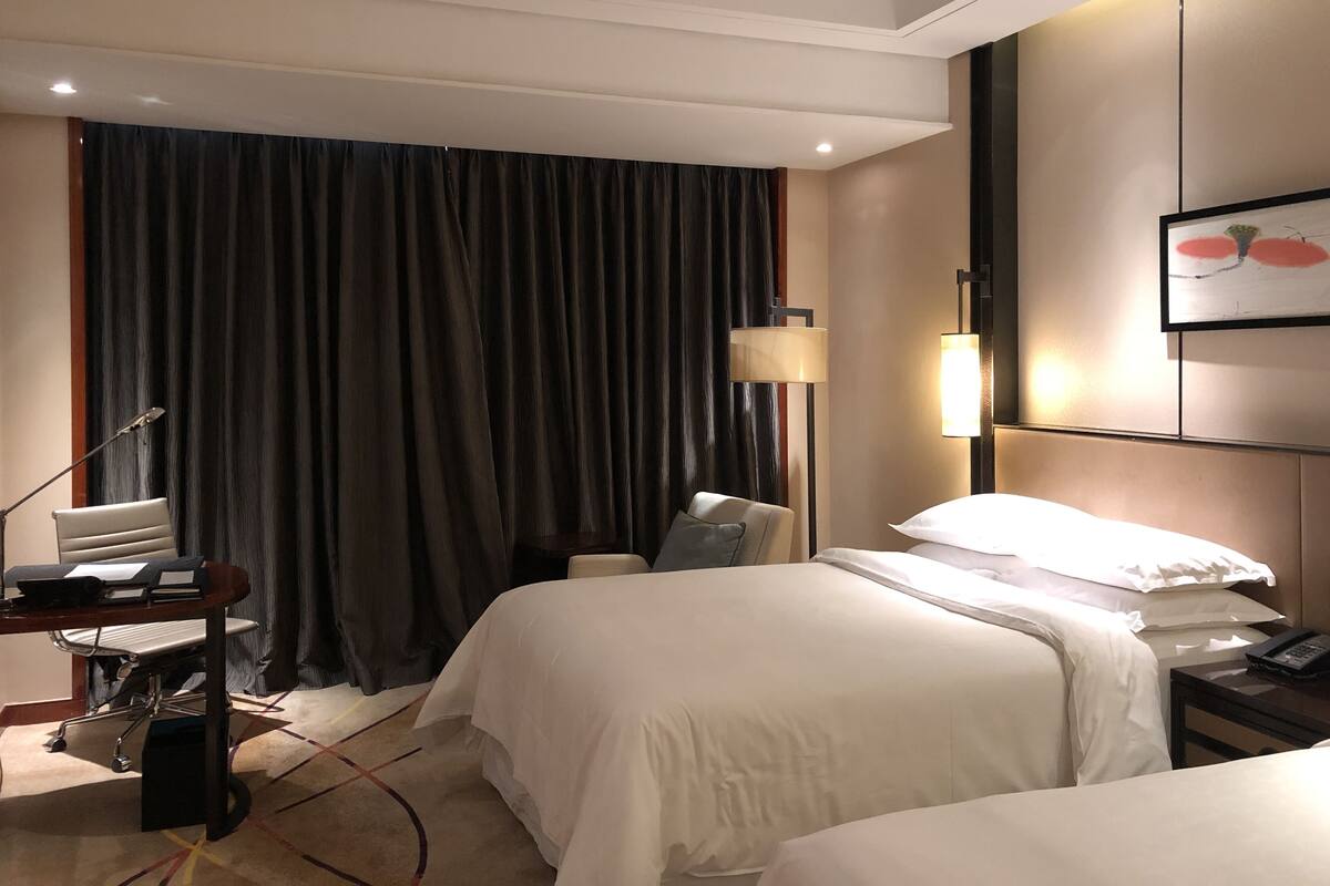 A hotel room with two beds, a desk with a chair, a lamp, and a painting on the wall.