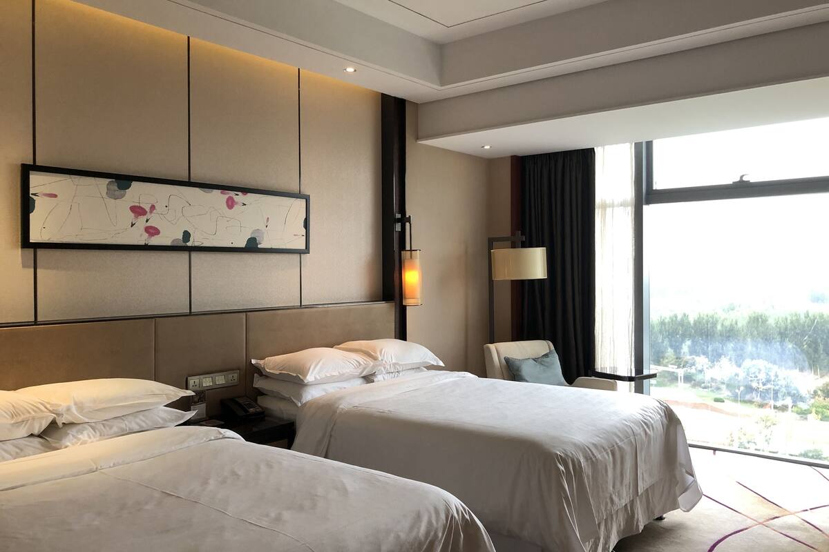 A hotel room with two beds, a large window, a painting on the wall, and a nightstand with a lamp.