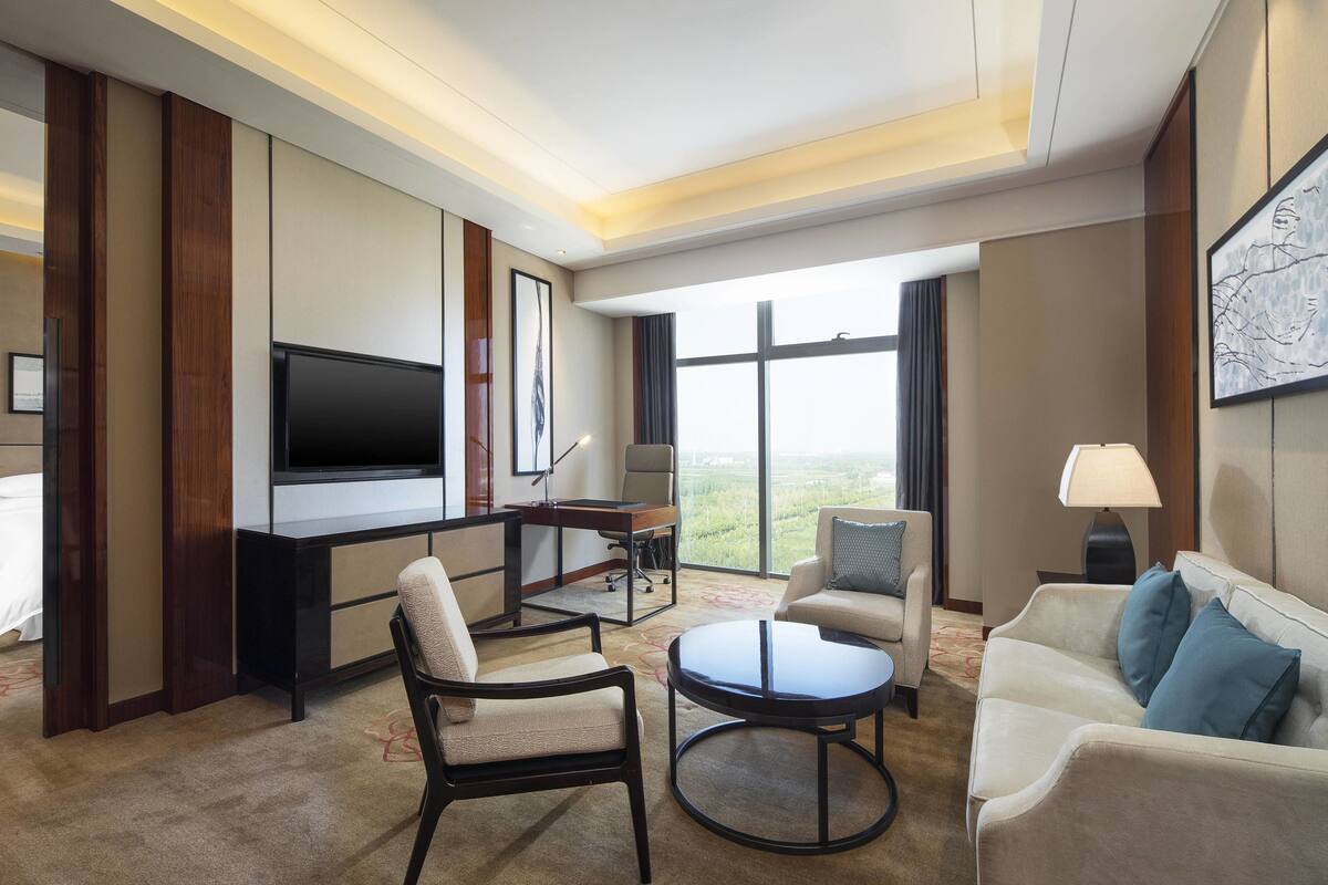 A hotel room with a flat-screen TV, a desk, two armchairs, a sofa, and a large window offering a view of the outdoors.