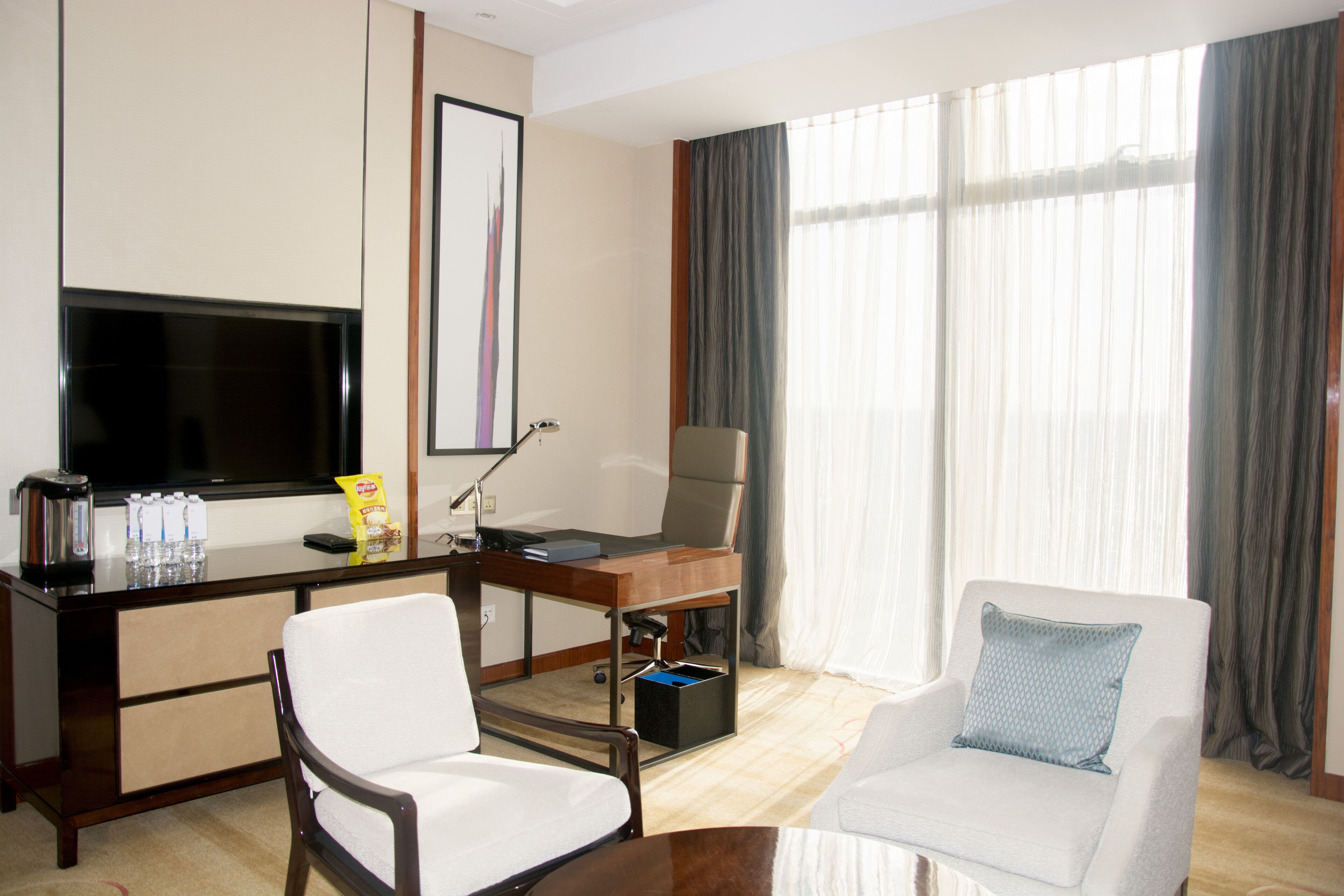 Hotelkamer met flatscreen televisie, bureau met stoel, bank en groot raam met gordijnen.