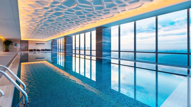 Una piscina interior moderna con fondo de cristal, grandes ventanales y un diseño geométrico en el techo.