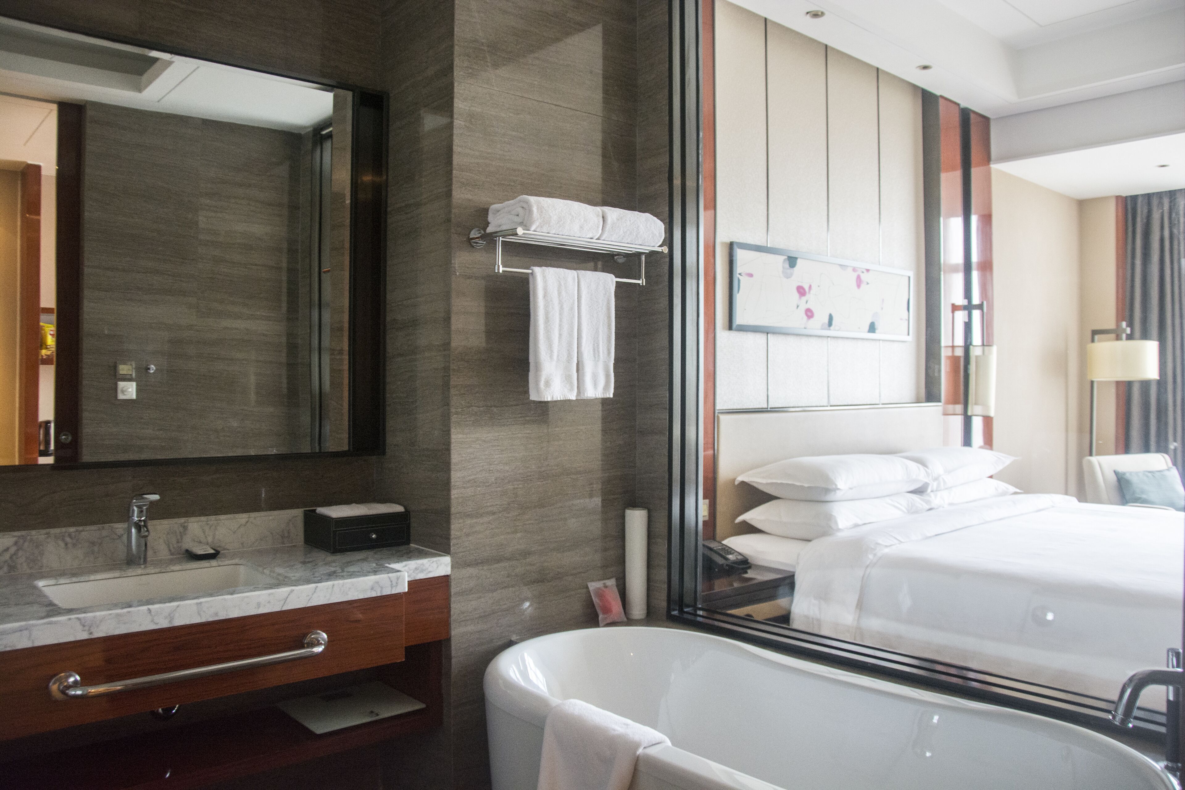A modern hotel bathroom with a large mirror, a marble sink, a bathtub, and a bed with white linens.