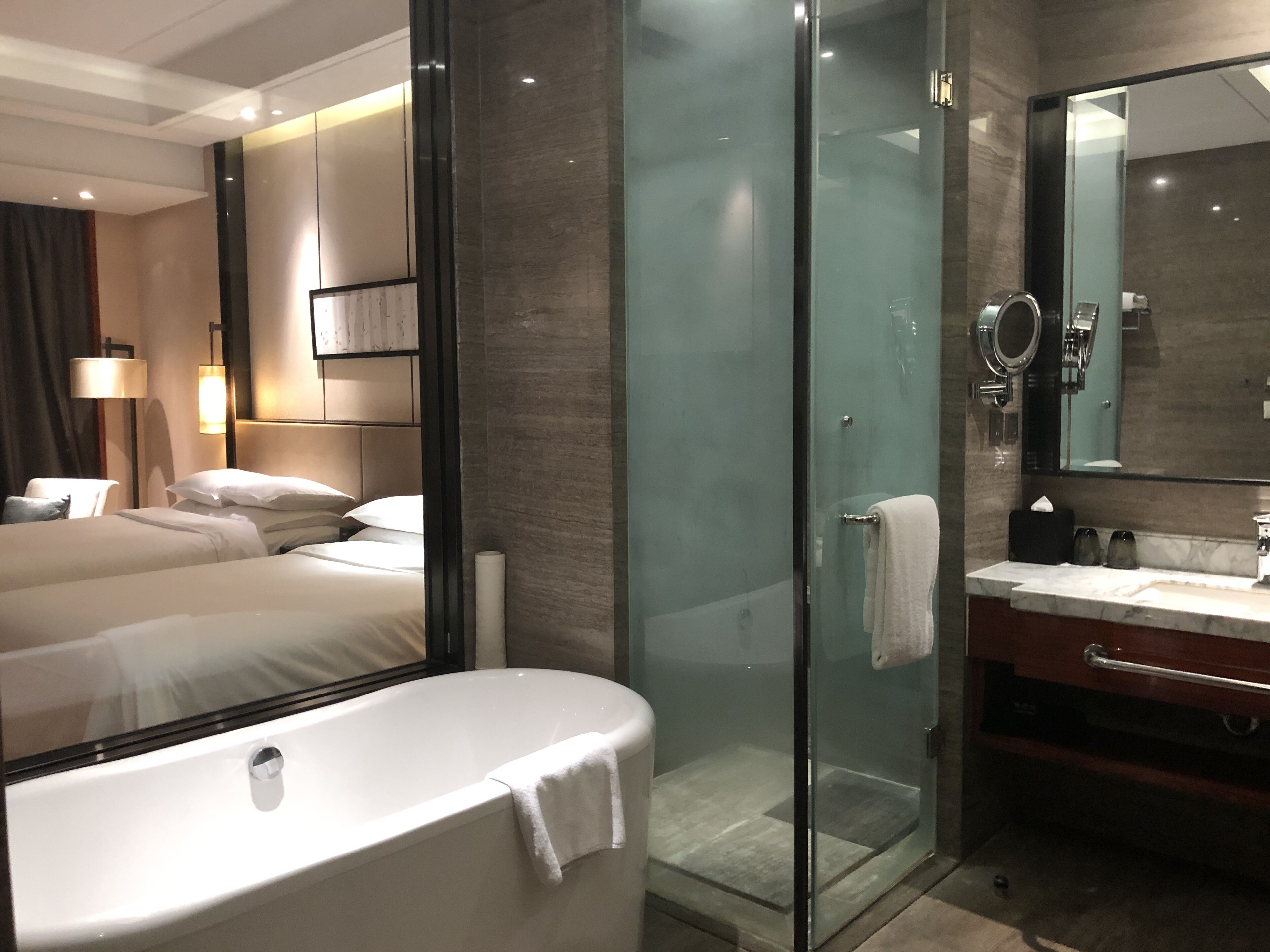 A modern hotel room with a glass-enclosed shower, a large bed with white linens, a vanity with a mirror, and a wall-mounted lamp.