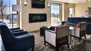 A hotel lobby with a flat-screen TV mounted on a brick wall, a seating area with blue upholstered chairs, a coffee table, and a large window offering a view of the parking lot. - Comfort Inn Midland South I-20 (Midland)