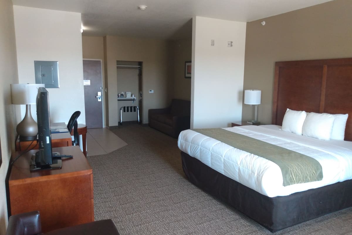 A hotel room with a large bed, a desk with a computer, a chair, a small kitchenette, and a door leading to another room.