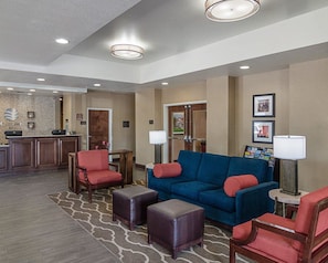 A hotel lobby with a reception desk, a blue sofa with red cushions, two red armchairs, a coffee table, and a patterned rug.