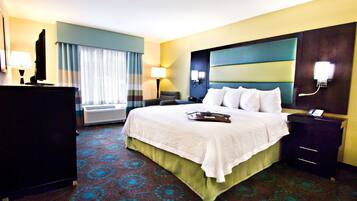 A hotel room with a large bed, a desk, a chair, a lamp, an air conditioning unit, and a nightstand with a phone.