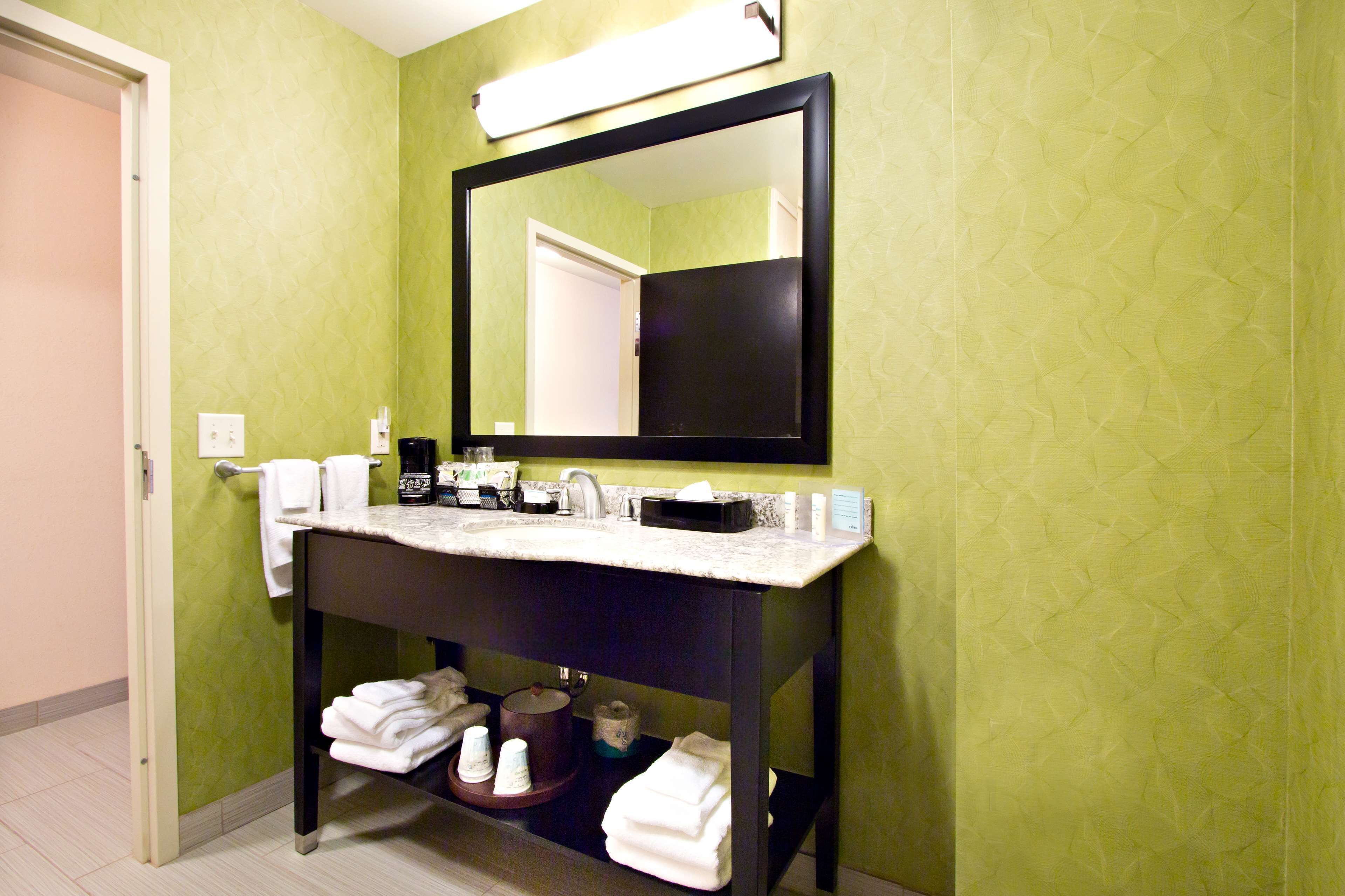 A bathroom with a green wall, a black-framed mirror, a white countertop, and a sink. There are towels and toiletries on the counter.