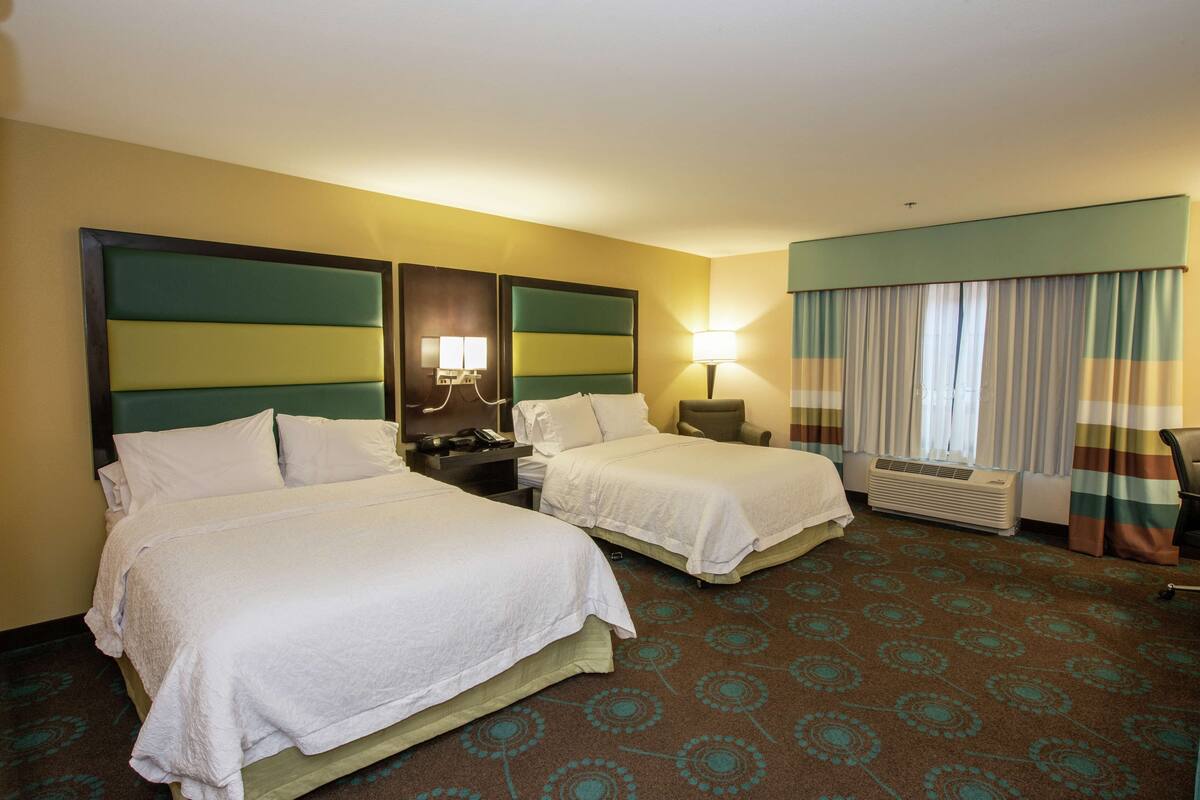 A hotel room with two beds, a nightstand, a lamp, a window with curtains, and a carpeted floor.