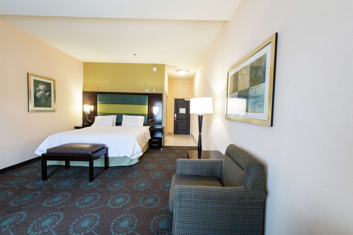 A hotel room with a bed, a desk, a chair, a lamp, and a painting on the wall.