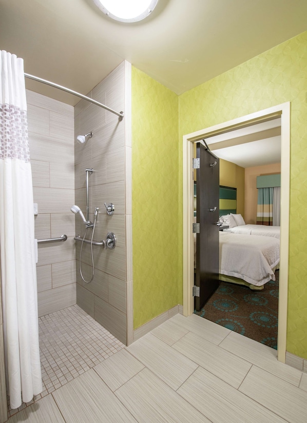 A hotel bathroom with a shower, toilet, and a bed visible through an open door.