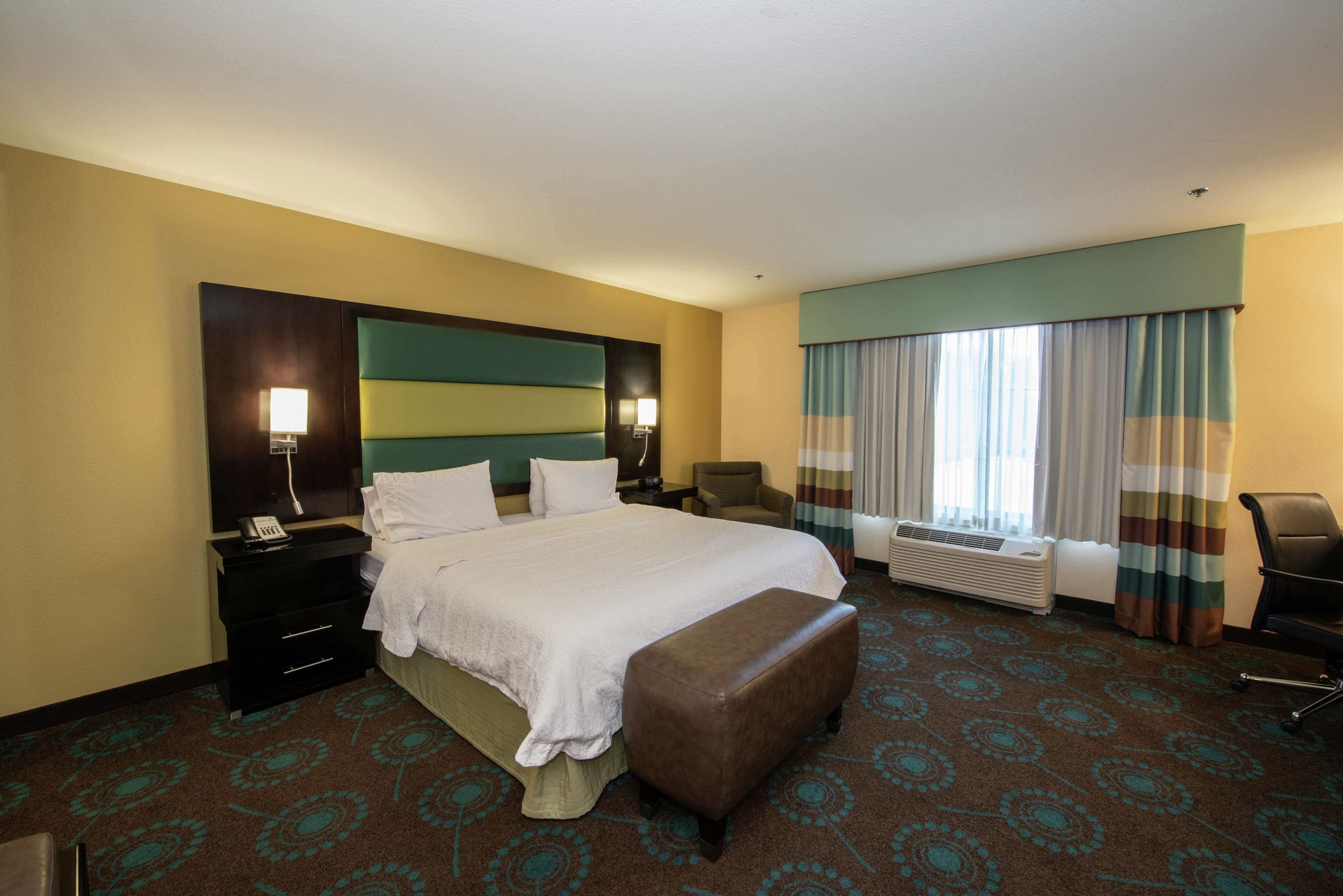 A hotel room with a large bed, a desk with a lamp, a chair, an air conditioning unit, and a window with curtains.