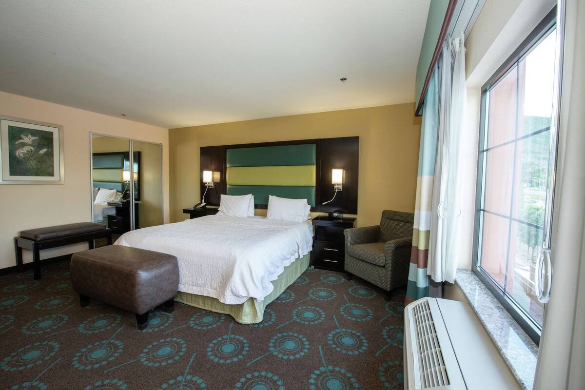 A hotel room with a large bed, a bench, a desk, and a window with curtains.