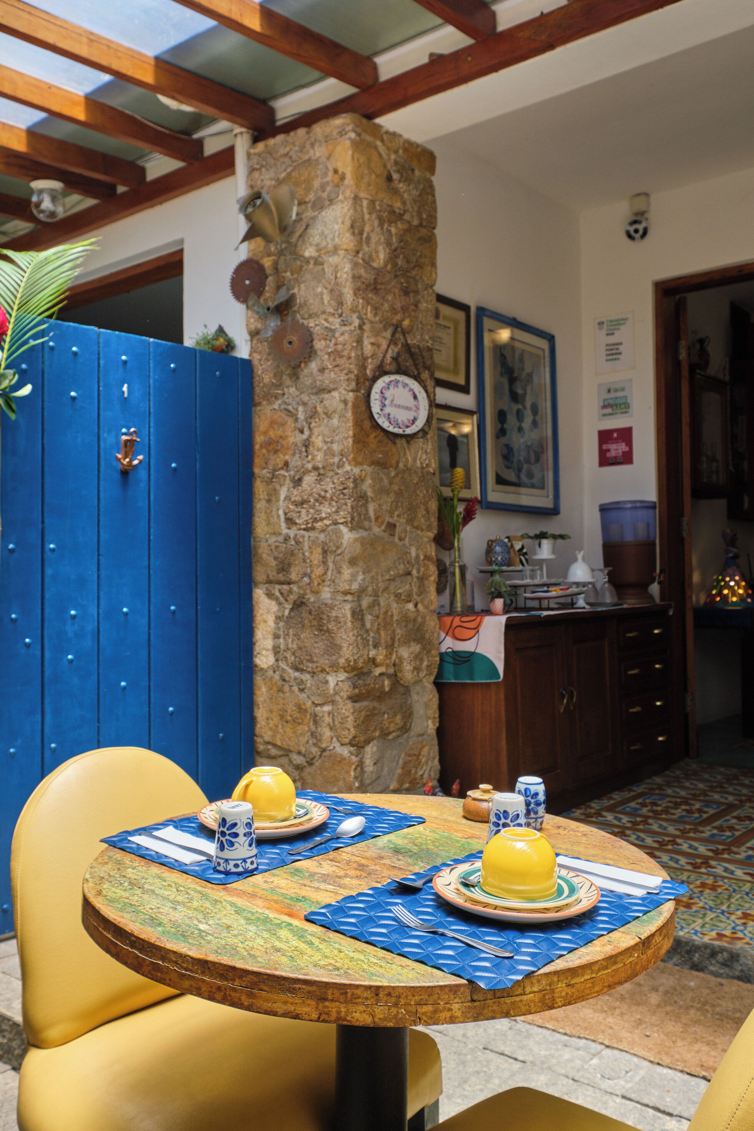 Un coin repas avec une table ronde dressée pour deux personnes, une porte bleue et un pilier en pierre.