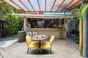 A covered outdoor dining area with a table and chairs, surrounded by tropical plants and a kitchen in the background. - Pousada Pontal Gardens (Paraty)