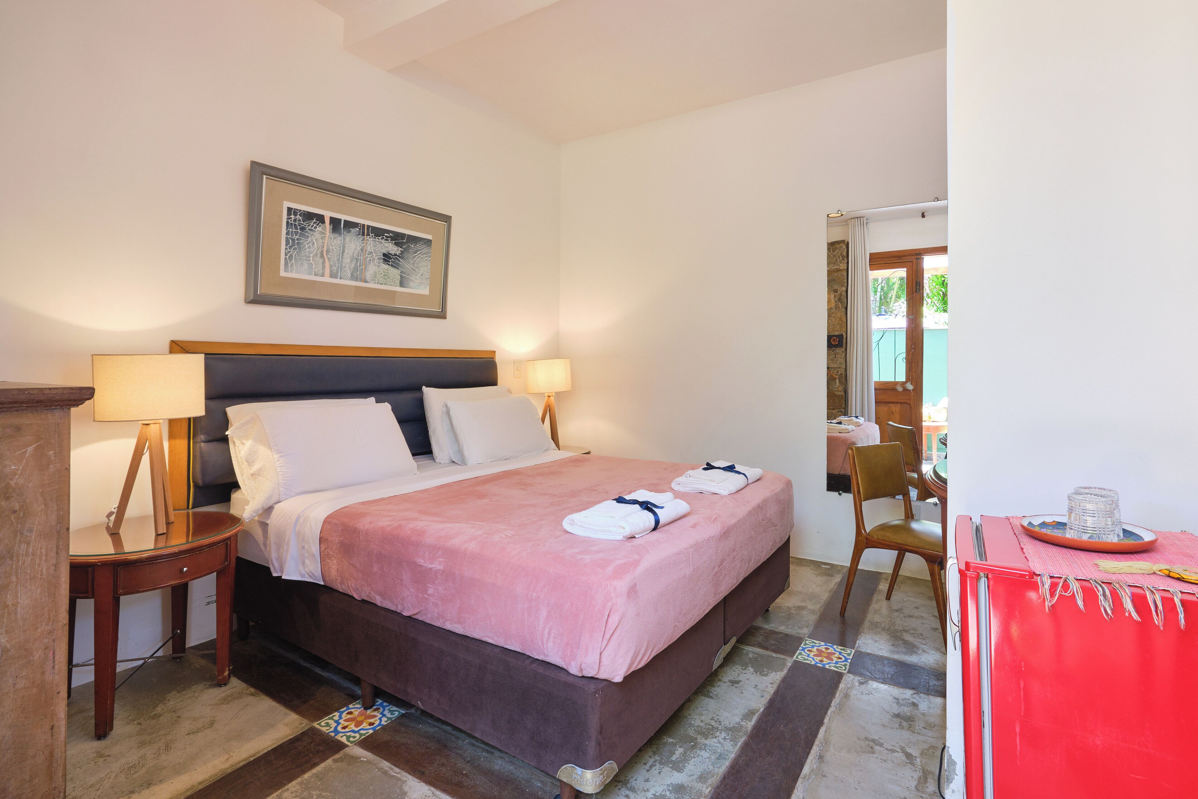 A neatly made bed with a pink bedspread and two towels, a wooden nightstand with a lamp, a framed picture on the wall, and a red cabinet with a tray.