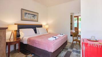 A neatly made bed with a pink bedspread and two towels, a wooden nightstand with a lamp, a framed picture on the wall, and a red cabinet with a tray.