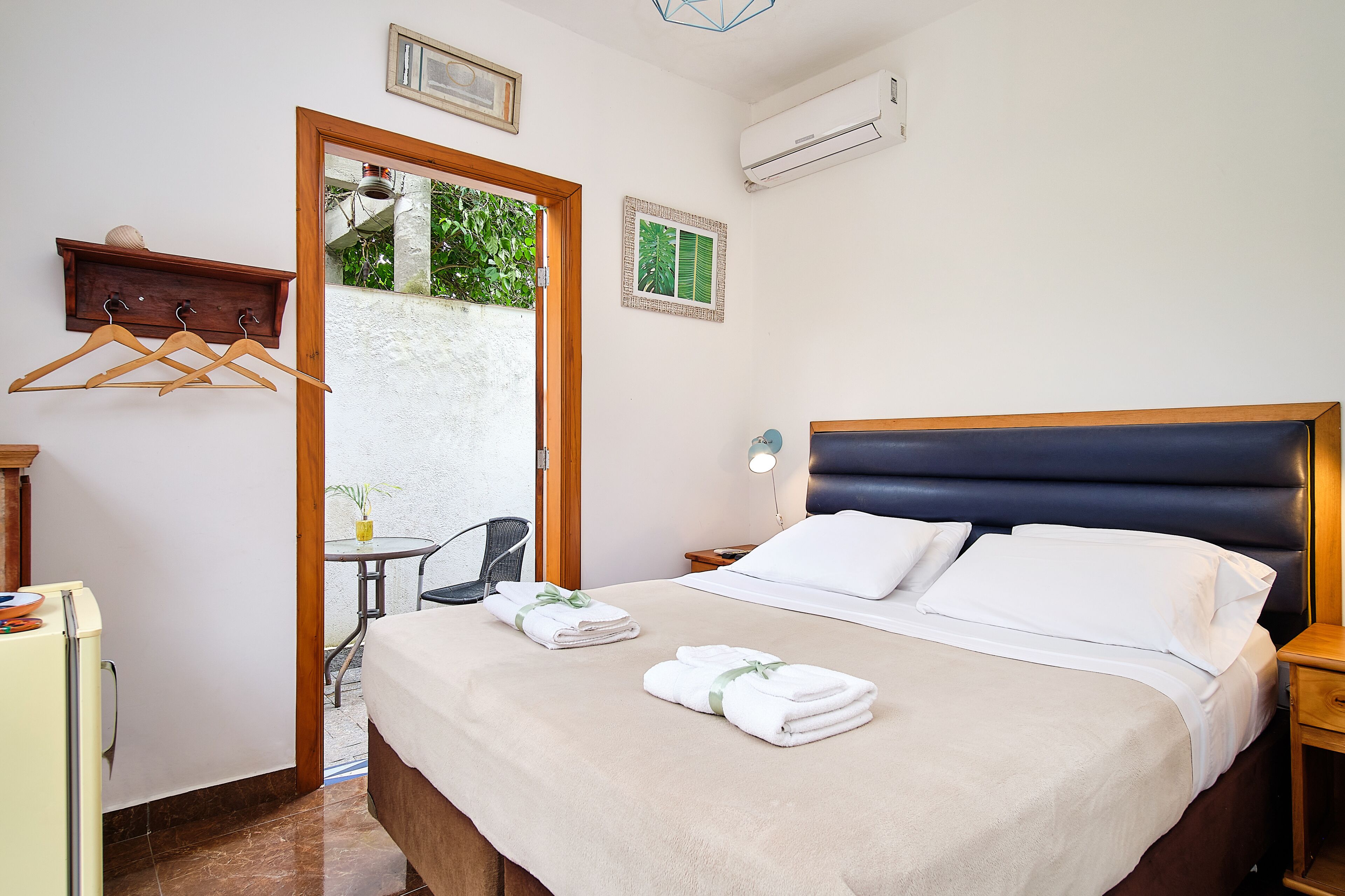 Une chambre d’hôtel comprenant un lit, des tables de chevet, un bureau, une chaise, un porte-manteau et une porte donnant sur un espace extérieur agrémenté de plantes.