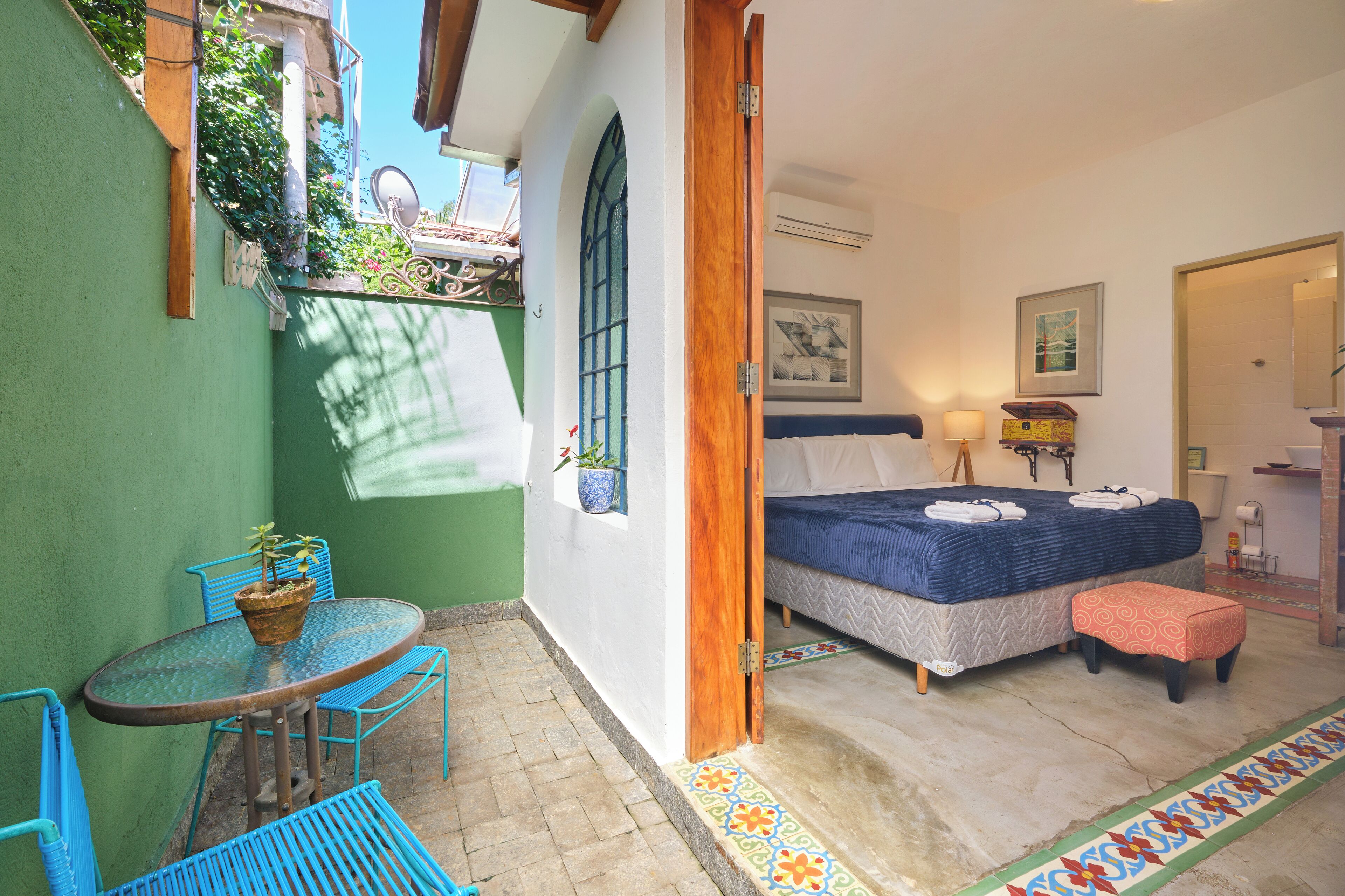 A room with a bed, a table with chairs, a small stool, a potted plant, and a window with a view of greenery.