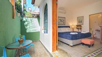 A room with a bed, a table with chairs, a small stool, a potted plant, and a window with a view of greenery.