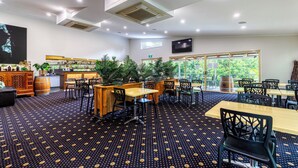 A restaurant with a patterned carpet, wooden tables and chairs, potted plants, and a television mounted on the wall. - Red Bridge Motor Inn (Woombye)