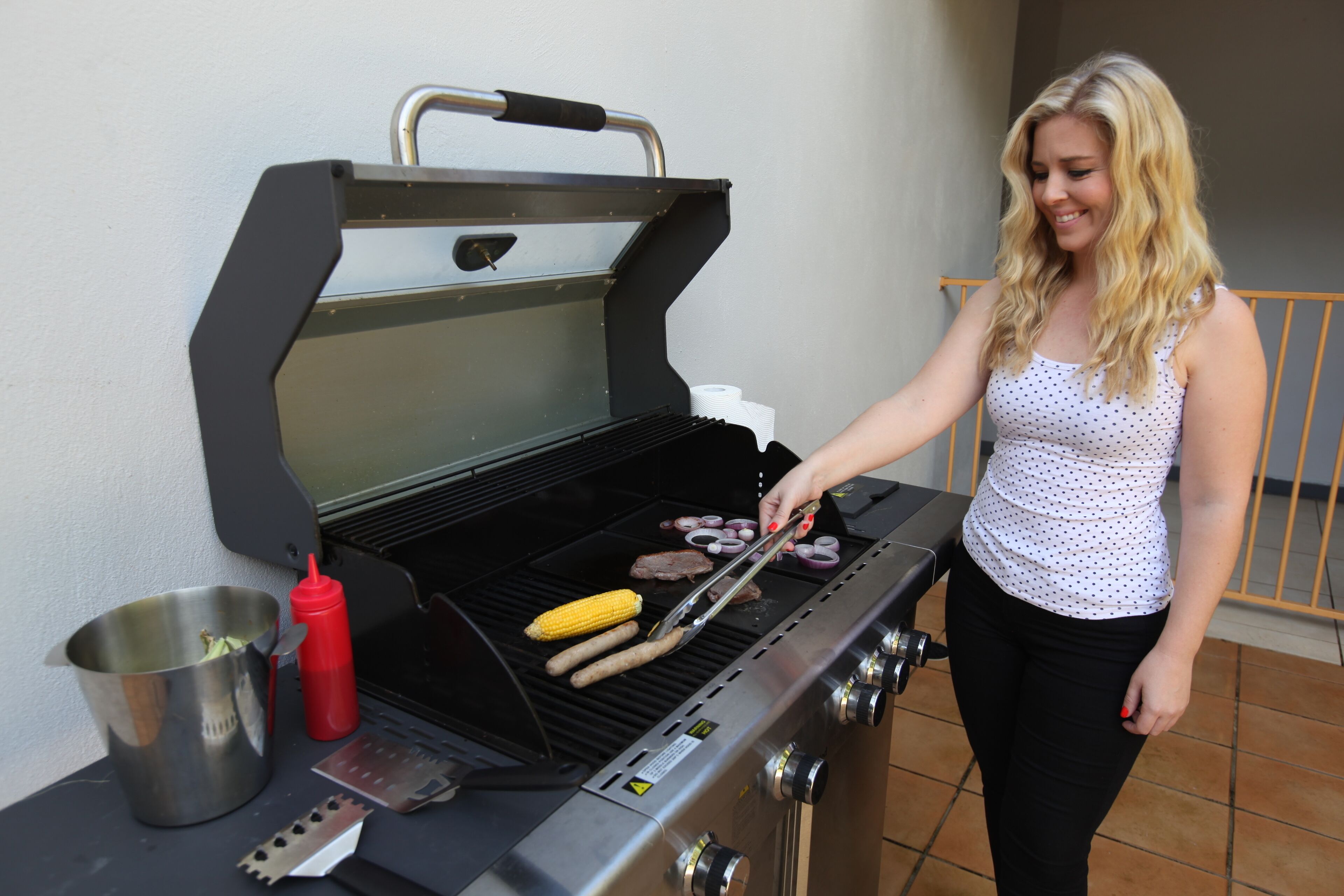 A woman grilling food on an outdoor grill with a metal pot and a bottle nearby.