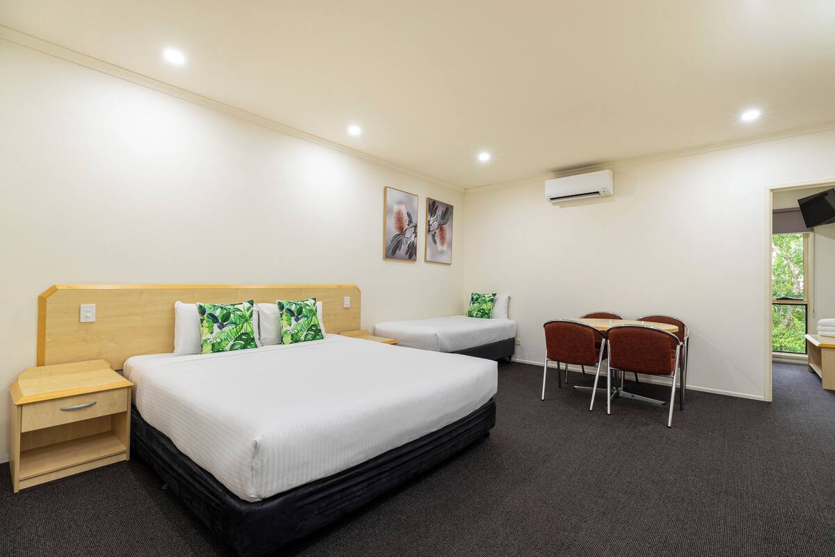 A hotel room with a large bed, two smaller beds, a desk with chairs, and a window.
