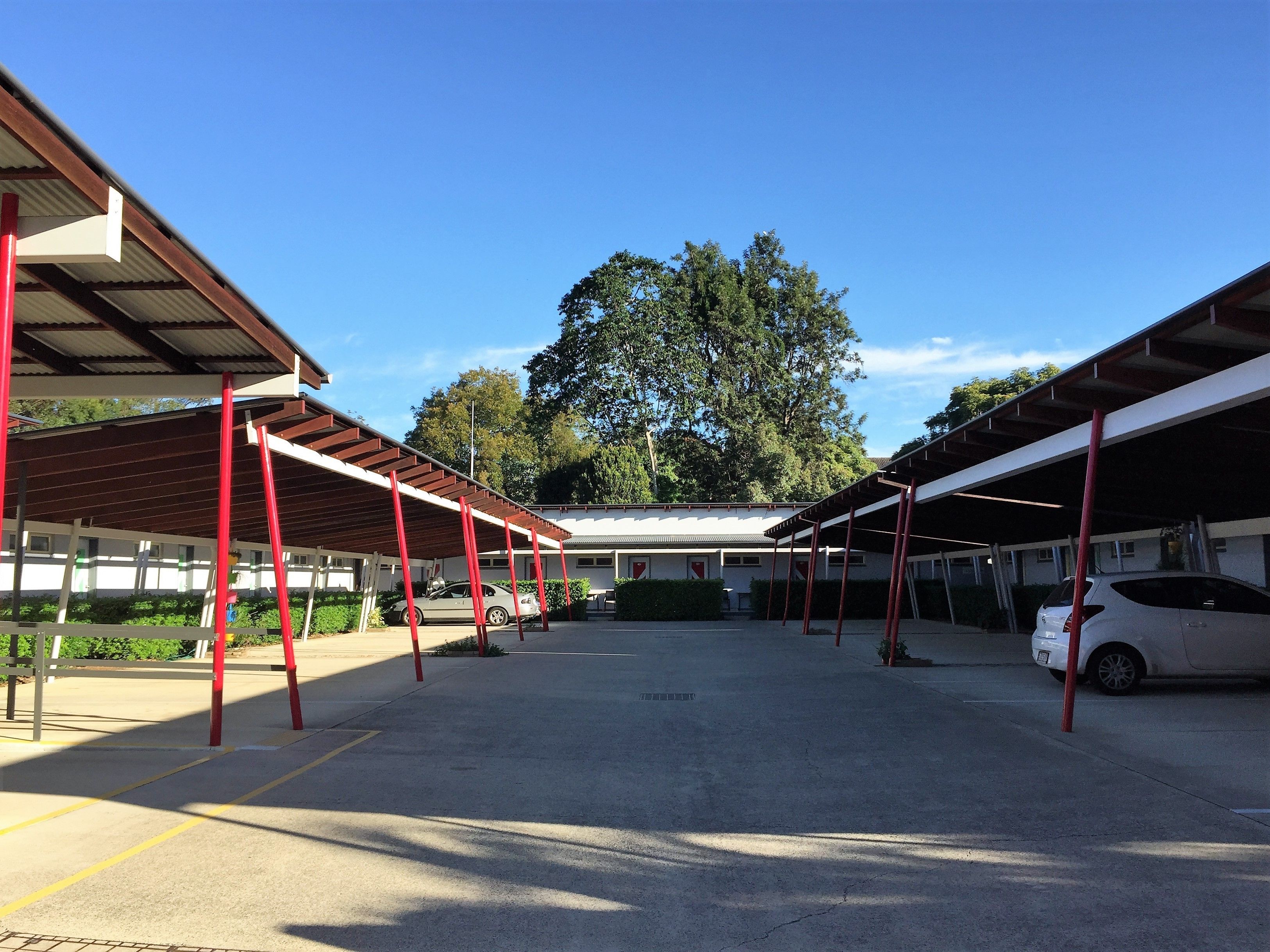 Uma área de estacionamento coberta com vários carros, um edifício com telhado plano e árvores ao fundo.