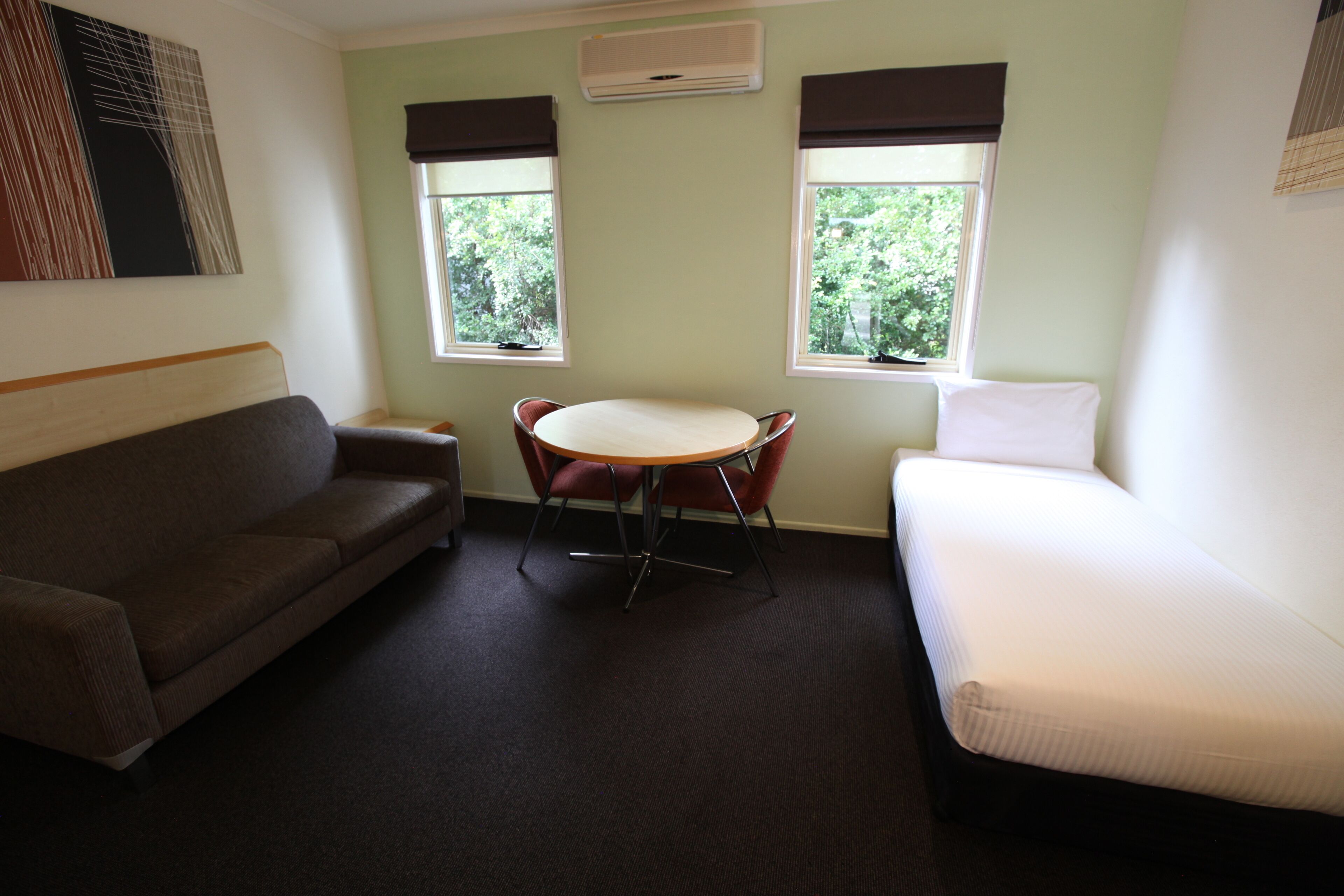 A room with a brown sofa, a round table with four chairs, and a white bed. There are two windows with brown curtains and an air conditioning unit above them.
