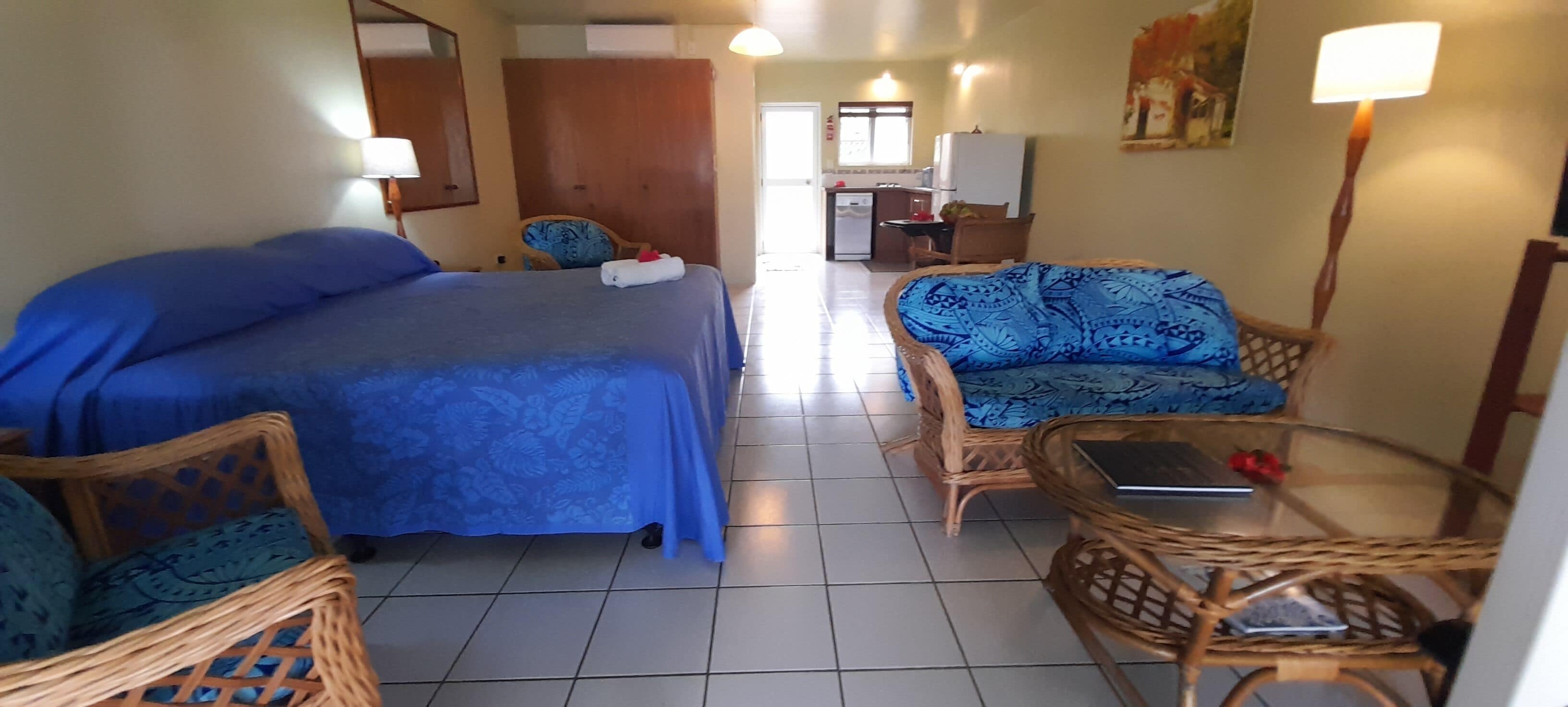 A hotel room with a bed, two wicker chairs, a small table, a desk, and a kitchen area in the background.