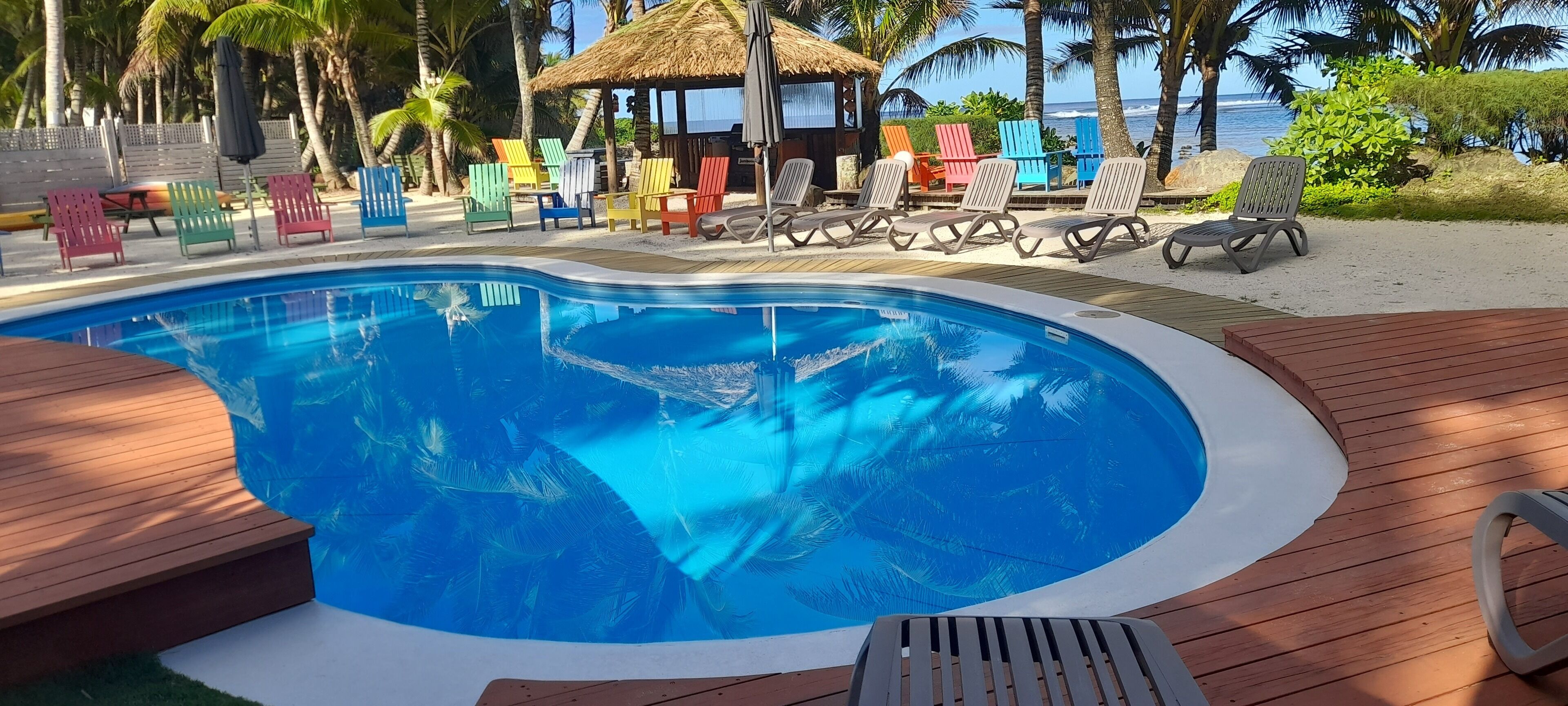 A pool area with a wooden deck, colorful lounge chairs, and a thatched-roof structure.
