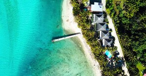 An aerial view of a beachside property with a pier extending into turquoise waters, surrounded by lush greenery and a row of buildings with sloped roofs. - Avana Waterfront Apartments (Rarotonga)