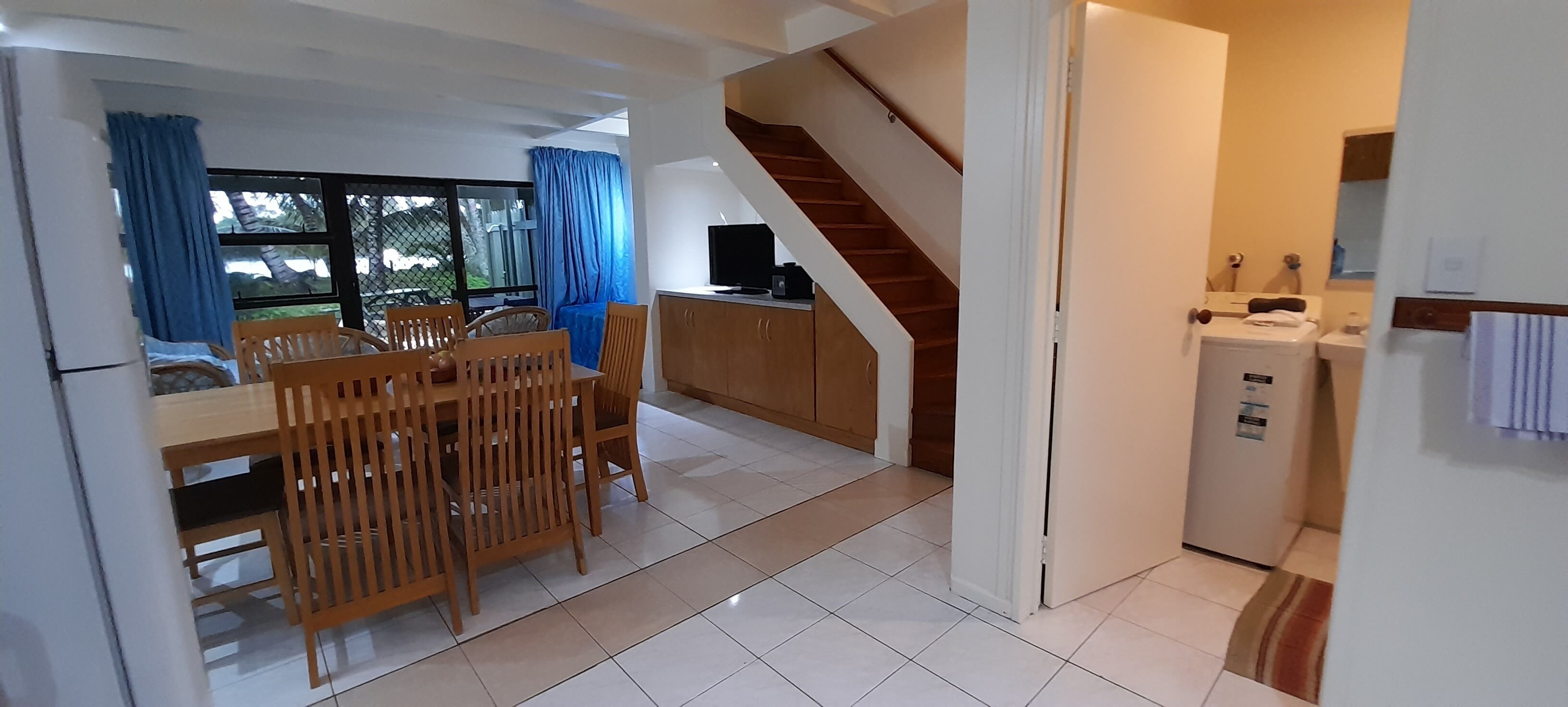 A modern kitchen with a built-in oven and a small refrigerator. There is a dining area with a wooden table and chairs, and a staircase leading to an upper level.
