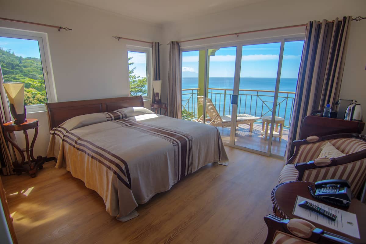 Quarto com cama grande, varanda e vista para o oceano, e área de estar com mesa e cadeiras.