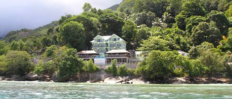 A multi-story house with a balcony, surrounded by lush greenery and a rocky hillside.