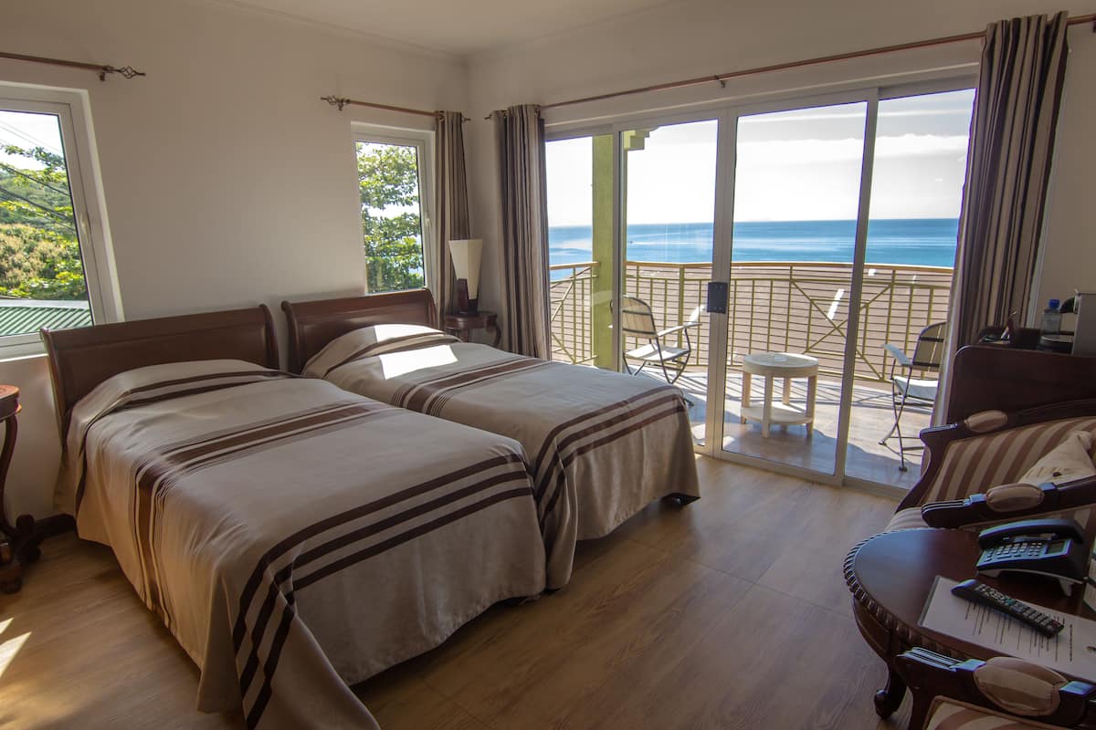 Quarto de hotel com duas camas, varanda com mesa e cadeiras, e escrivaninha com telefone e controles remotos.