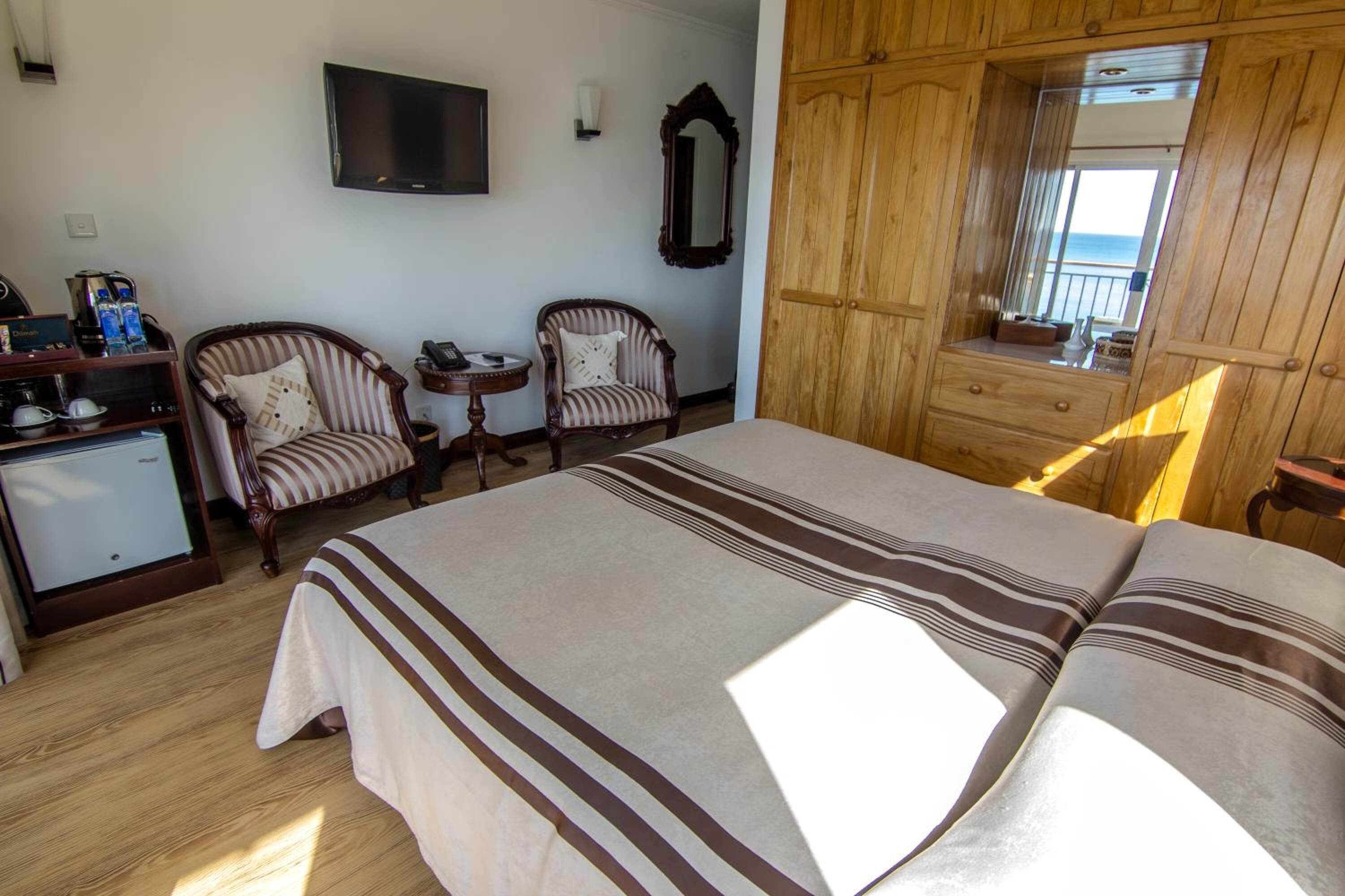 A hotel room with a bed, two armchairs, a small table, a desk with a computer, a TV, and a wooden wardrobe.