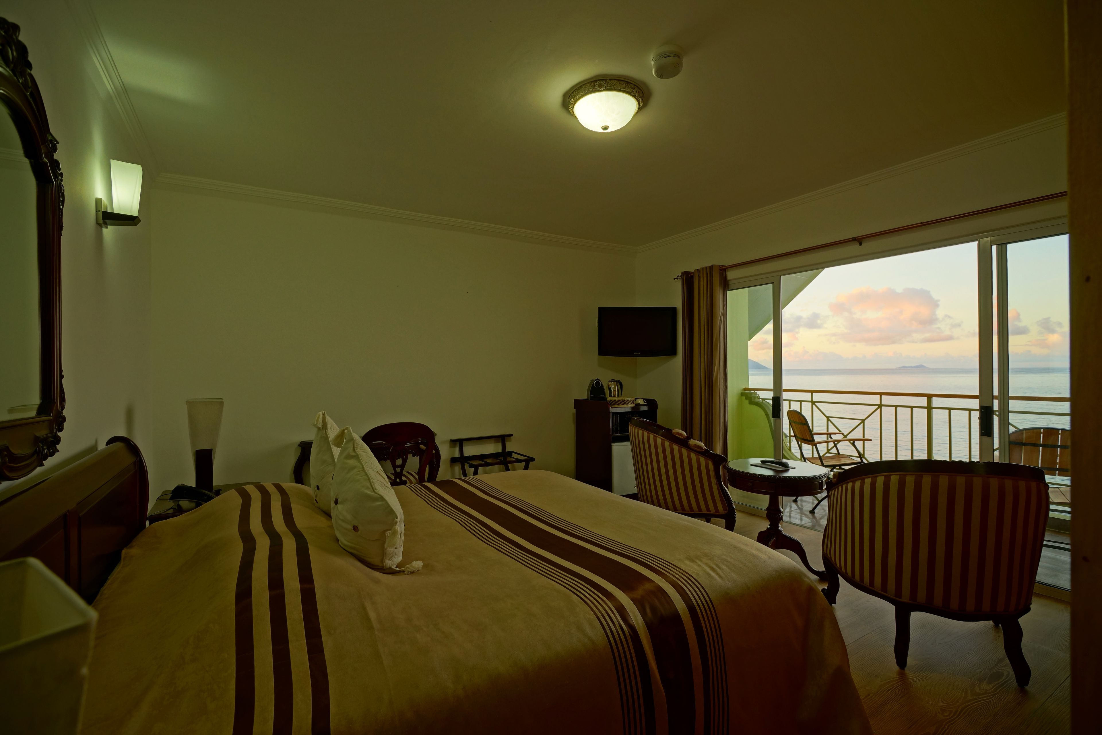 A hotel room with a bed, a chair, a small table, a TV, and a balcony with a view of the sea.