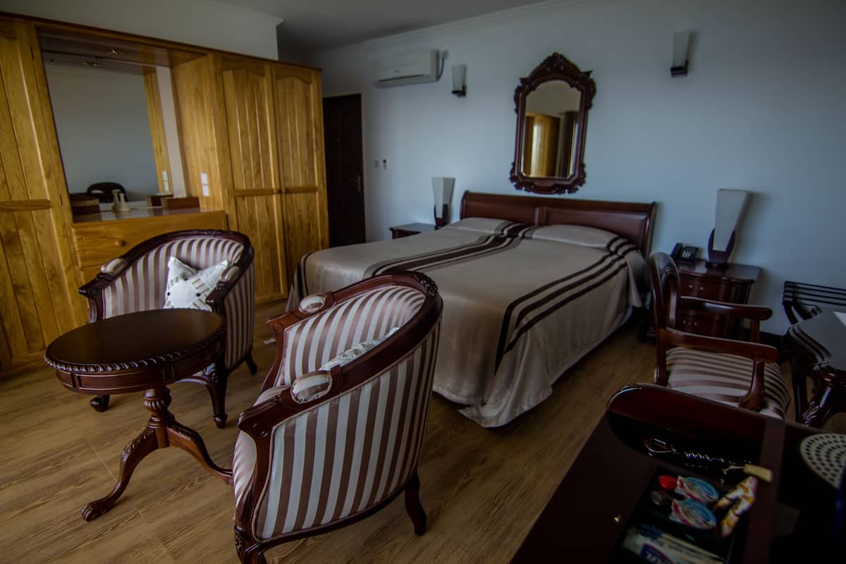 Quarto de hotel com uma cama grande, duas mesas de cabeceira, um guarda-roupa de madeira, uma poltrona listrada e uma pequena mesa redonda.
