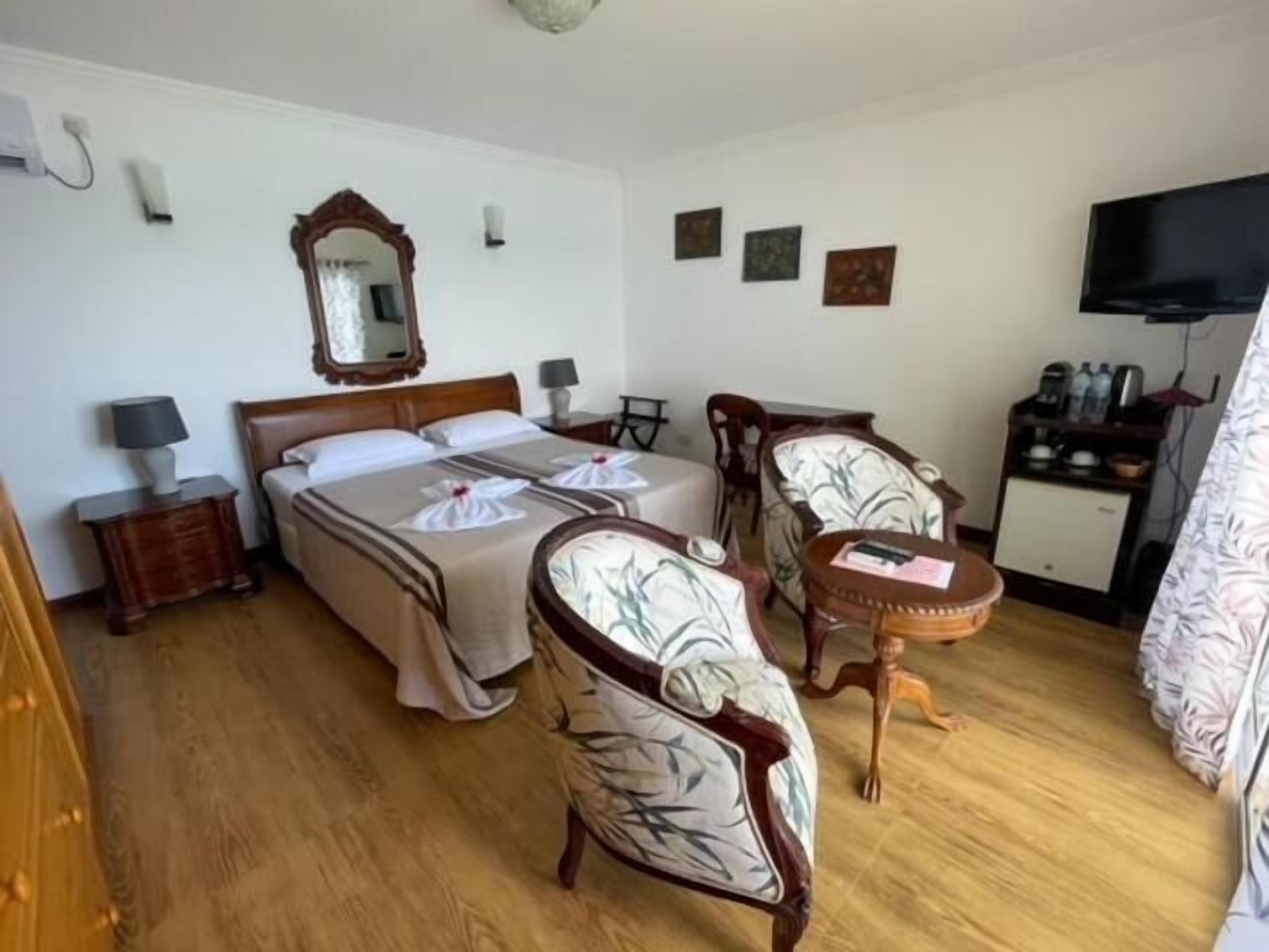 A hotel room with a bed, two chairs, a small table, a TV, and a minibar.